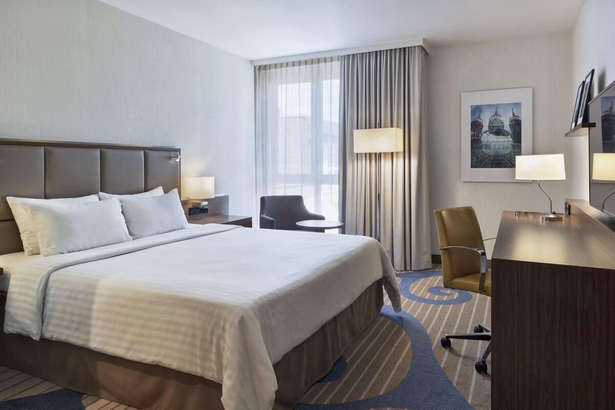 Ein Hotelzimmer mit einem großen Bett, einem Schreibtisch, einem Stuhl, einer Lampe und einem Fenster mit Vorhängen.