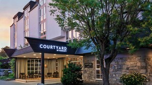 A Courtyard by Marriott hotel entrance with a covered outdoor seating area, stone facade, and a large tree in the foreground. - Courtyard by Marriott Wiesbaden-Nordenstadt (Wiesbaden)