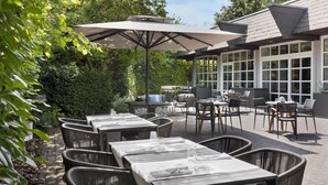 An outdoor dining area with wicker chairs and tables, large umbrellas, and a view of the surrounding greenery. - Courtyard by Marriott Wiesbaden-Nordenstadt (Wiesbaden)
