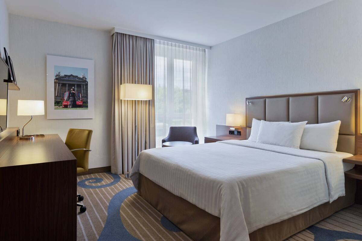 Ein Hotelzimmer mit einem großen Bett, einem Schreibtisch, einem Stuhl, einer Lampe und einem gerahmten Bild an der Wand.