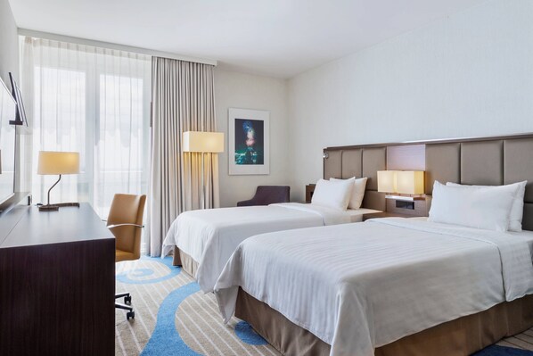 A hotel room with two beds, a desk, a chair, and a large window with curtains.