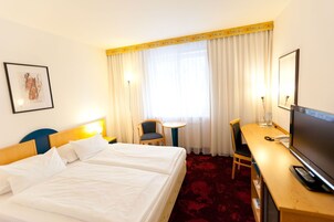 Quarto de hotel com duas camas, uma escrivaninha com televisão, uma cadeira, uma mesinha e uma janela com cortinas.