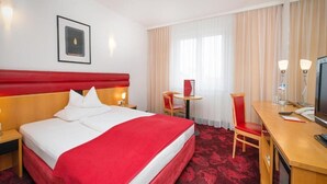 Ein Hotelzimmer mit einem großen Bett, einem Schreibtisch, einem Sessel, einem Fernseher und einem Fenster mit Vorhängen.
