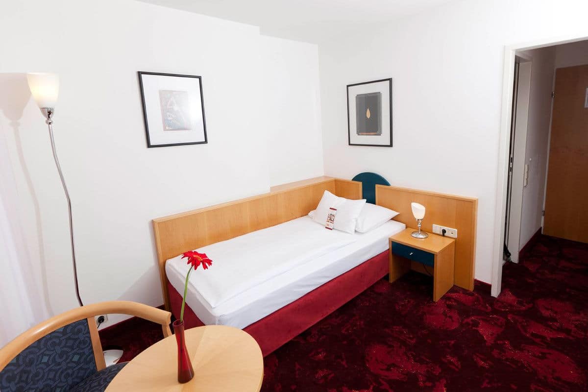 A hotel room with a single bed, a bedside table with a lamp, a chair, and a red carpet.