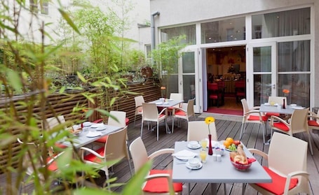 An outdoor dining area with tables and chairs, a wooden deck, and a view of the interior dining room through glass doors.
