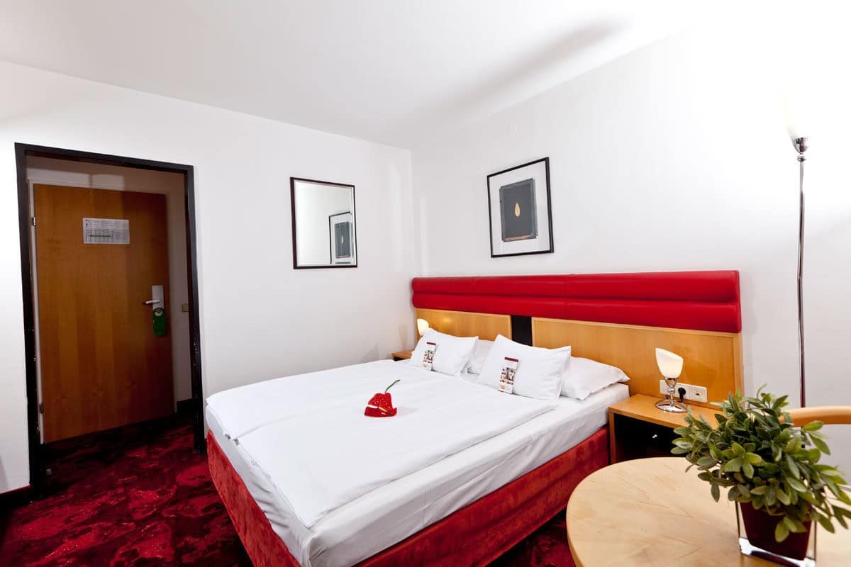 A hotel room with a bed, a bedside table with a lamp, a small table, and a potted plant.