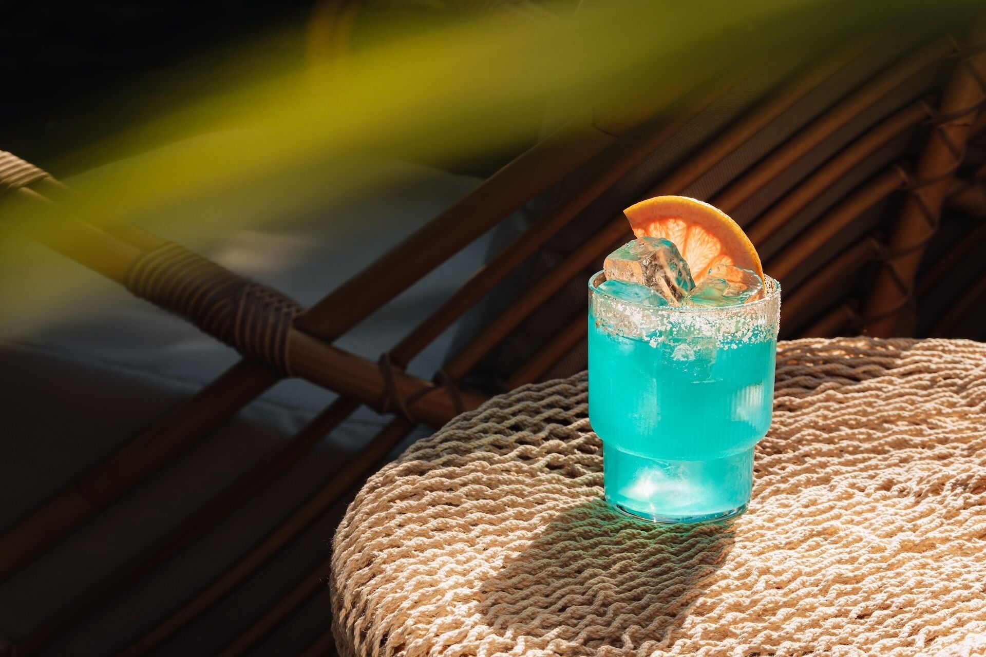 A glass of blue beverage with an orange slice garnish on a woven chair.