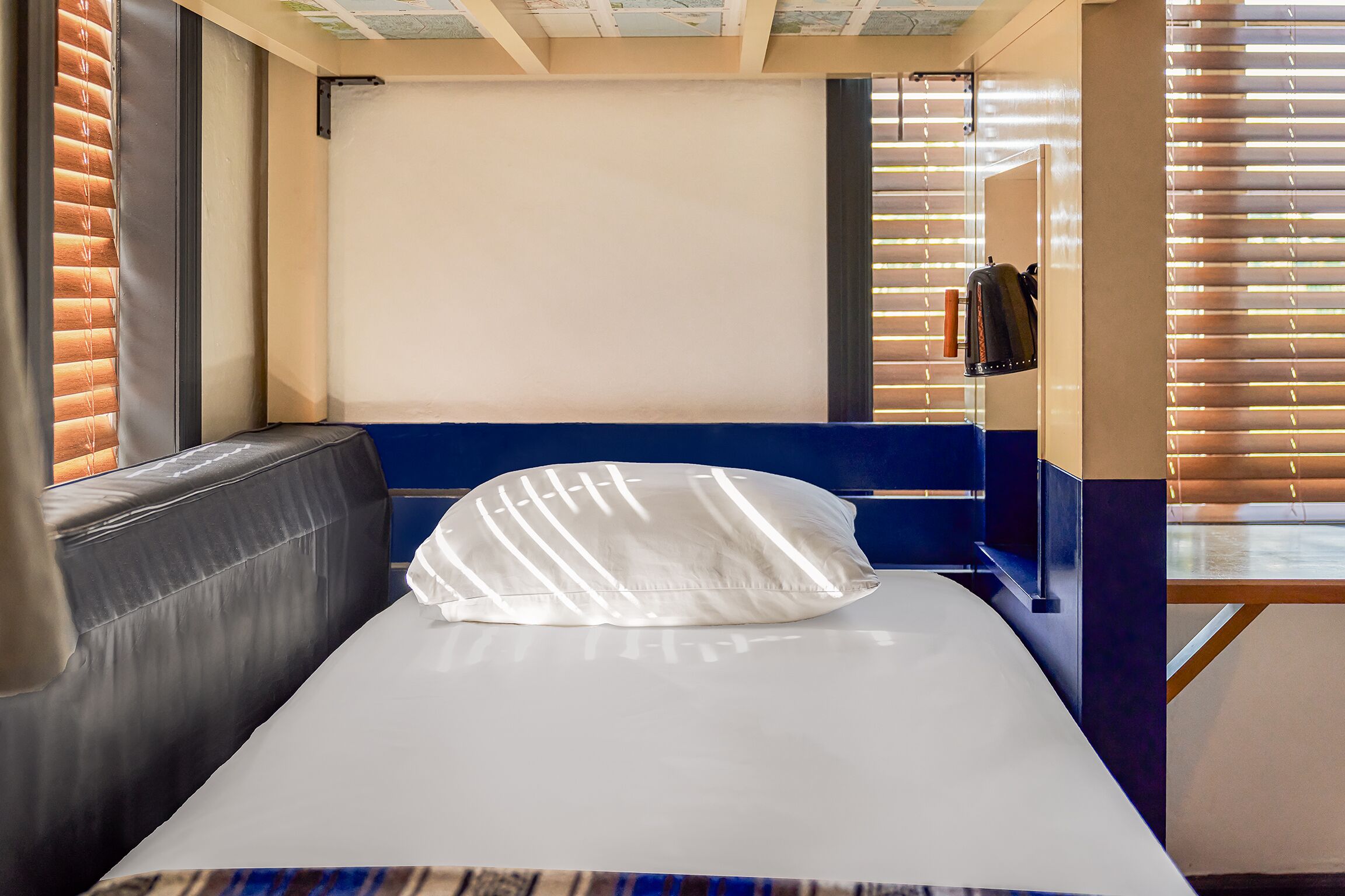 Ein Einzelbett mit weißer Matratze und Kissen, ein blauer Kopfteil sowie ein Fenster mit Jalousien.