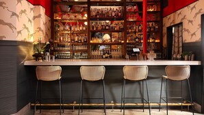 Una zona de bar con tres taburetes de mimbre, una barra con botellas y estantes llenos de licores.