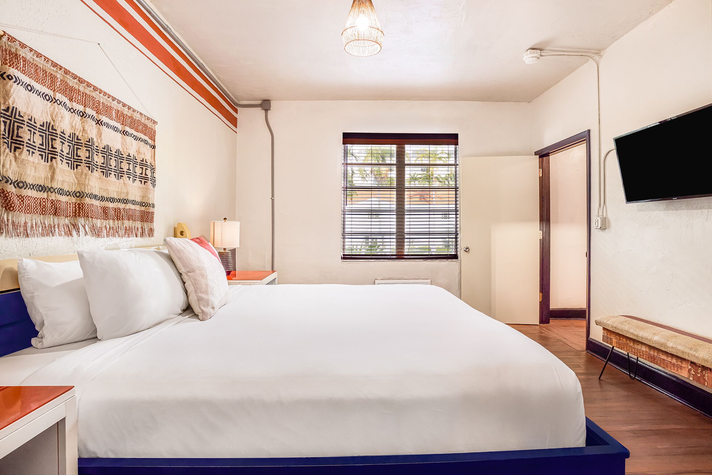 A hotel room with a large bed, a flat-screen TV mounted on the wall, a window with blinds, and a patterned tapestry on the wall.
