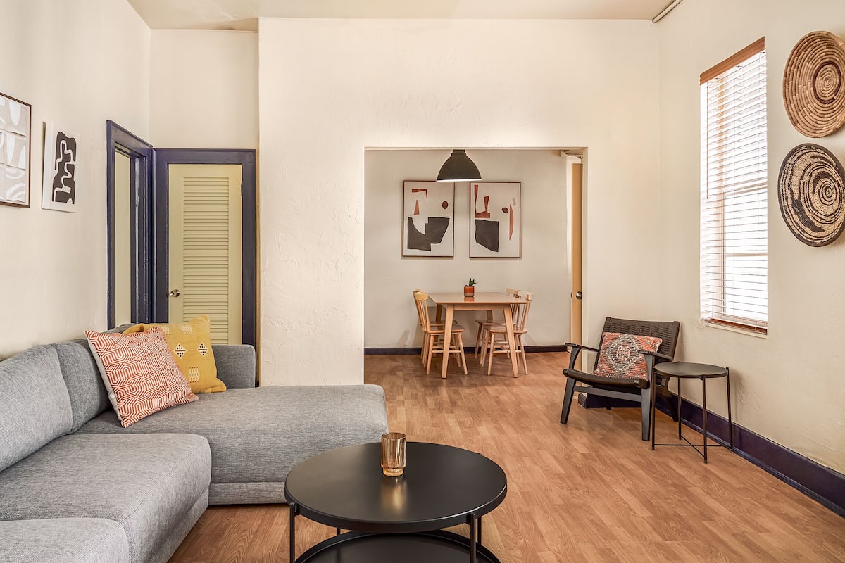 A living room with a gray sofa, a round black coffee table, and a dining area with a wooden table and chairs.
