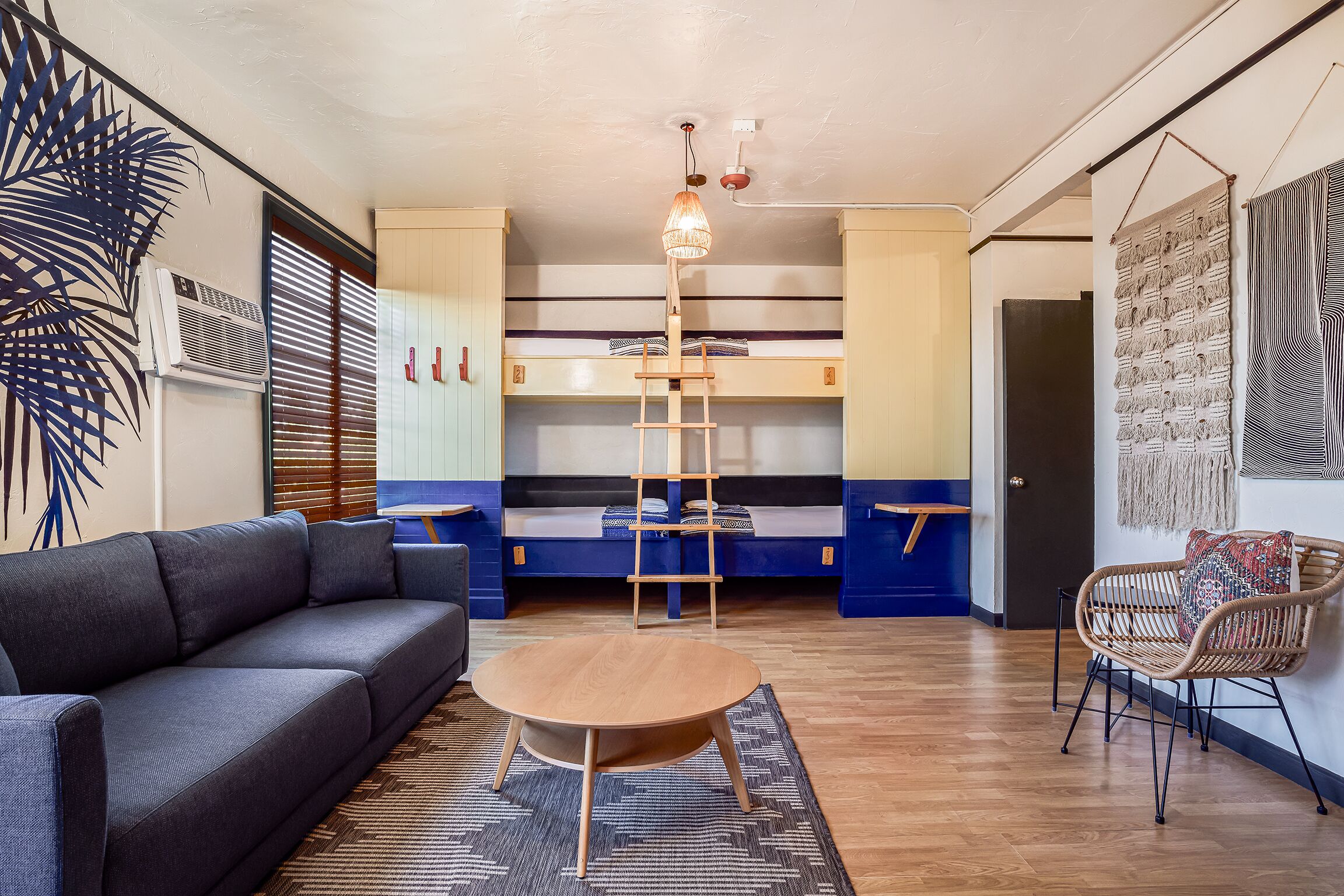 A modern living room with a sofa, a round wooden table, a ladder, bunk beds, and a wall-mounted macrame piece.