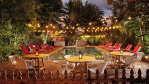 Zona de piscina exterior con sillones y mesas, rodeada de follaje e iluminación con luces colgantes.