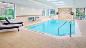 An indoor swimming pool with lounge chairs and tables, a stone wall, and large windows. - TownePlace Suites by Marriott Mississauga-Arpt Corp Ctr (Mississauga)