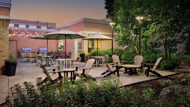 An outdoor patio area with tables and chairs, surrounded by greenery and a building in the background. - TownePlace Suites by Marriott Mississauga-Arpt Corp Ctr (Mississauga)
