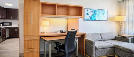 A hotel room with a kitchenette, a desk, a sofa, and a painting on the wall.