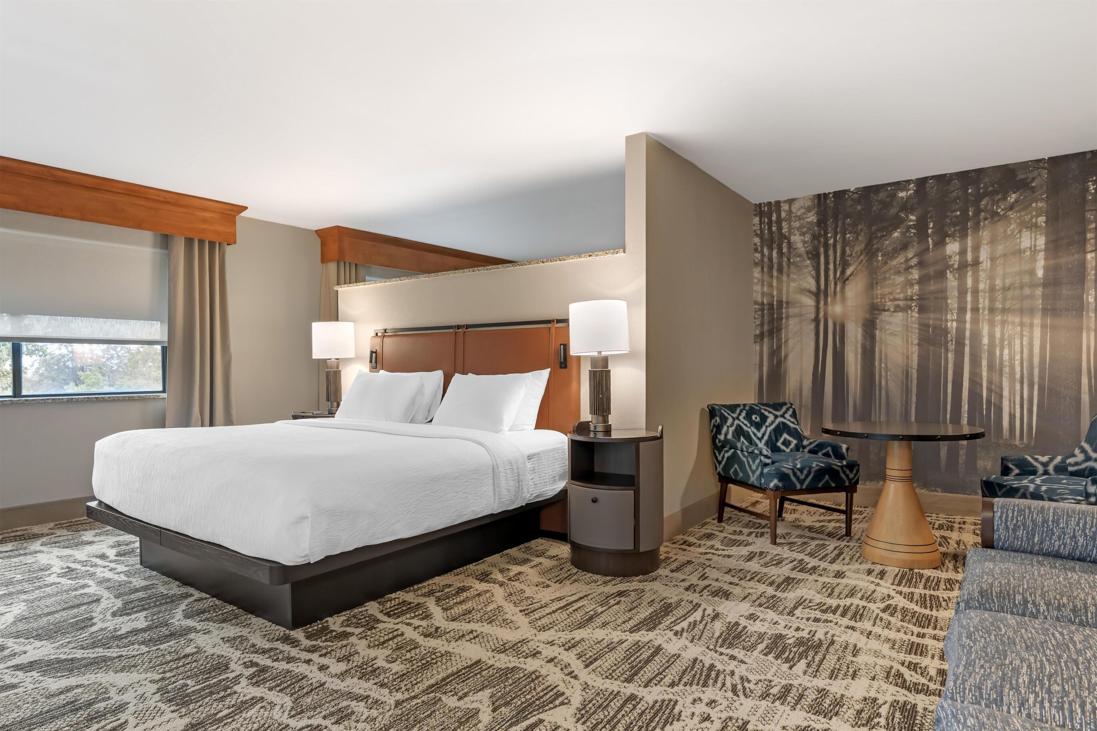 A hotel room with a large bed, two bedside tables with lamps, a chair, a round table, and a large window.