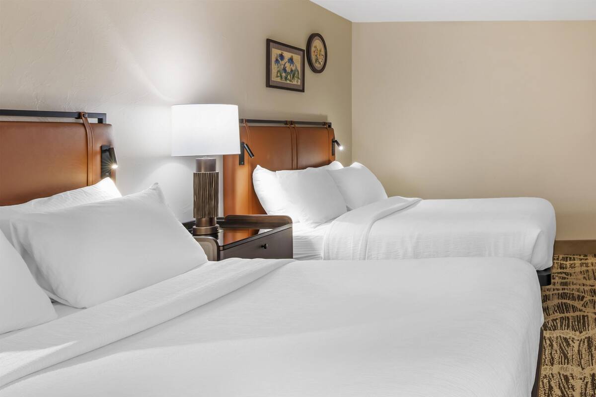 A hotel room with two beds, a nightstand, a lamp, and a wall clock.