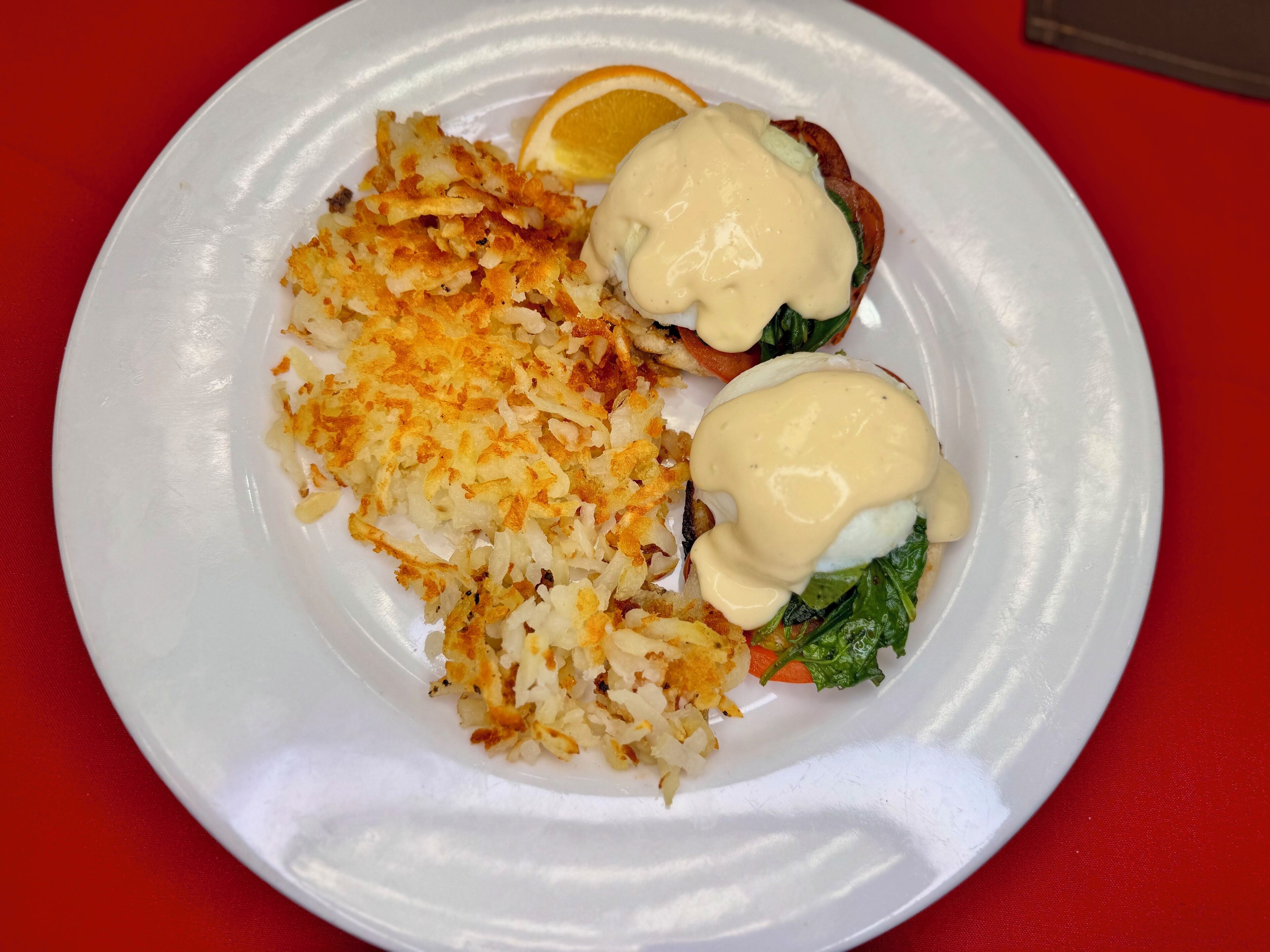 Une assiette blanche avec deux sandwichs œufs Bénédicte, des pommes de terre rissolées et une tranche d’orange.
