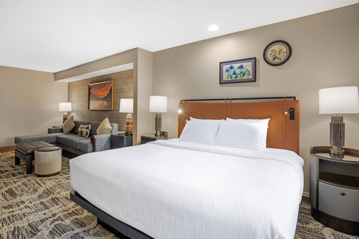 A hotel room with a large bed, a sofa, two side tables with lamps, a wall-mounted clock, and a framed picture.
