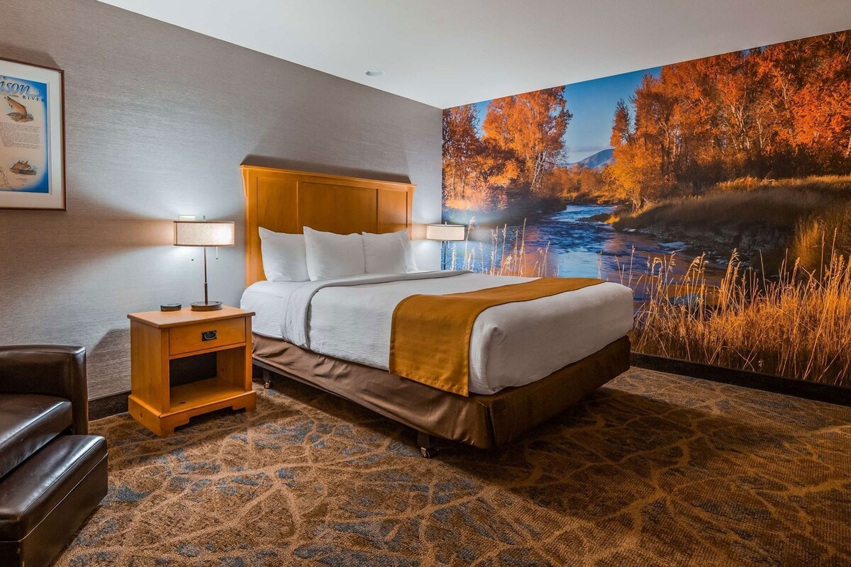 A hotel room with a bed, nightstand, lamp, chair, and a large wall mural.