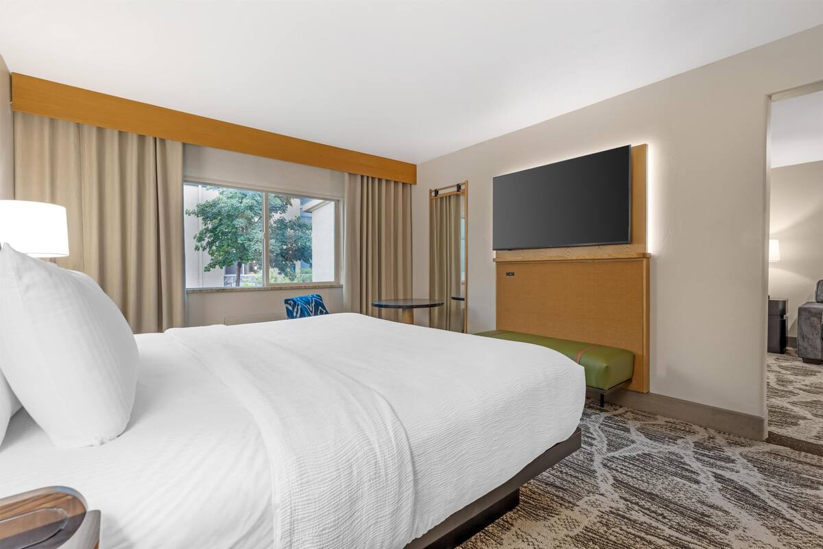 A hotel room with a large bed, a flat-screen TV mounted on the wall, a small table, and a green bench.