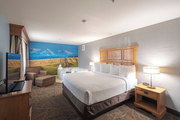 A hotel room with a large bed, a desk with a computer, a chair, a small table, and a painting of a mountain landscape on the wall.