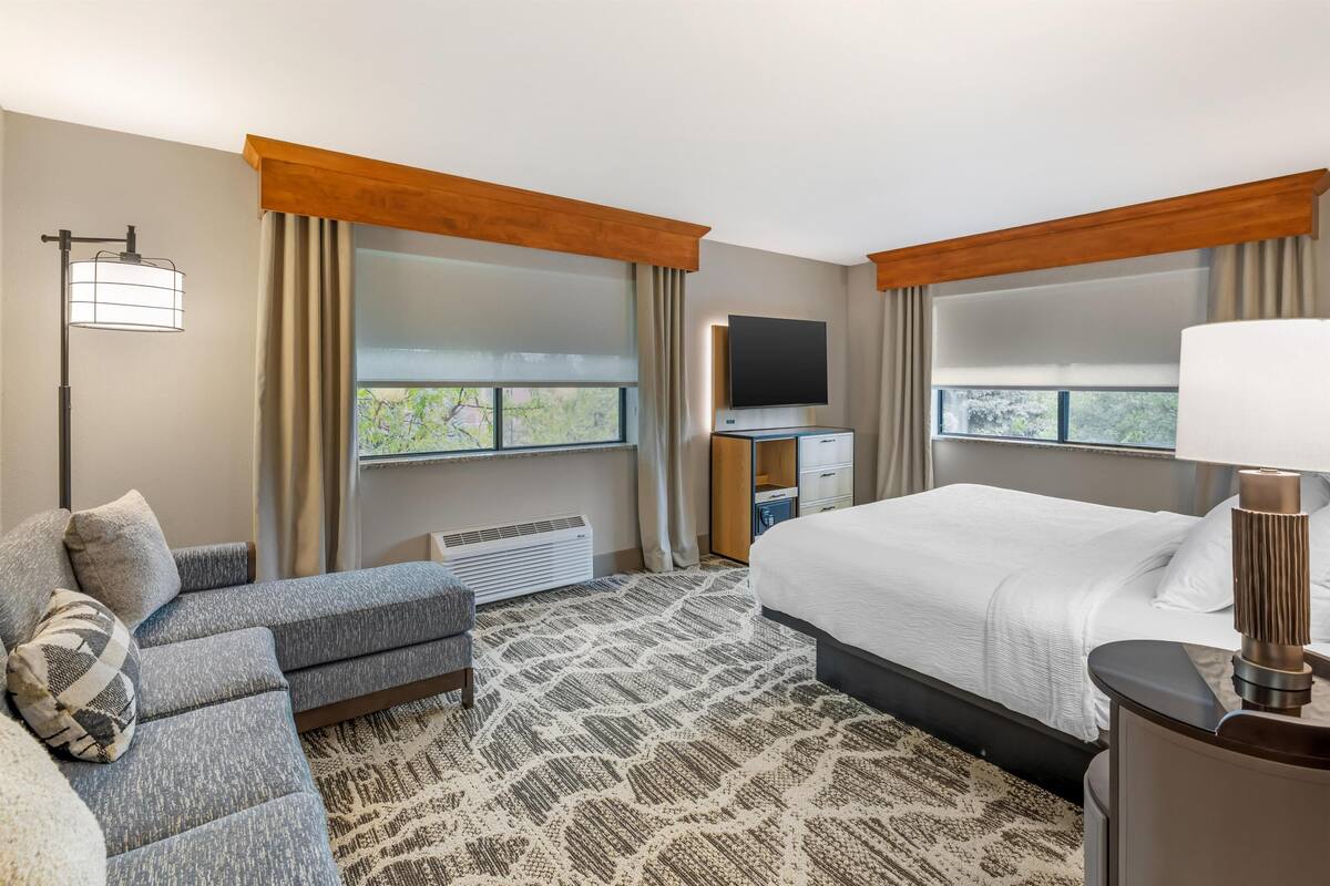 Une chambre d’hôtel avec un lit, un canapé, une télévision, un bureau et une fenêtre avec des rideaux.