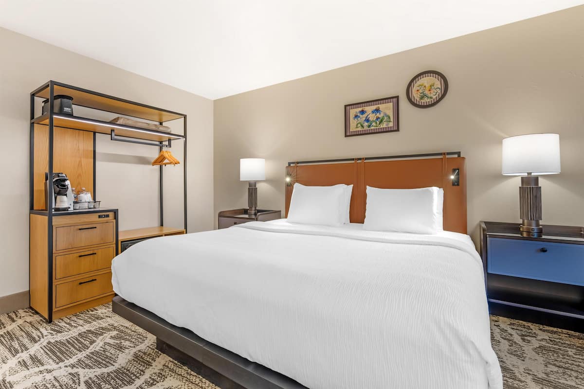 A hotel room with a bed, a wooden wardrobe, a desk, two bedside tables with lamps, and a wall clock.