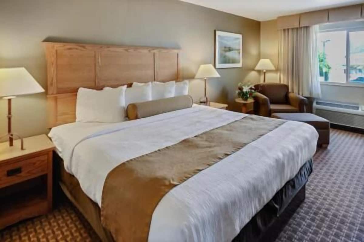A hotel room with a large bed, two bedside tables, a chair, a window with curtains, and a painting on the wall.