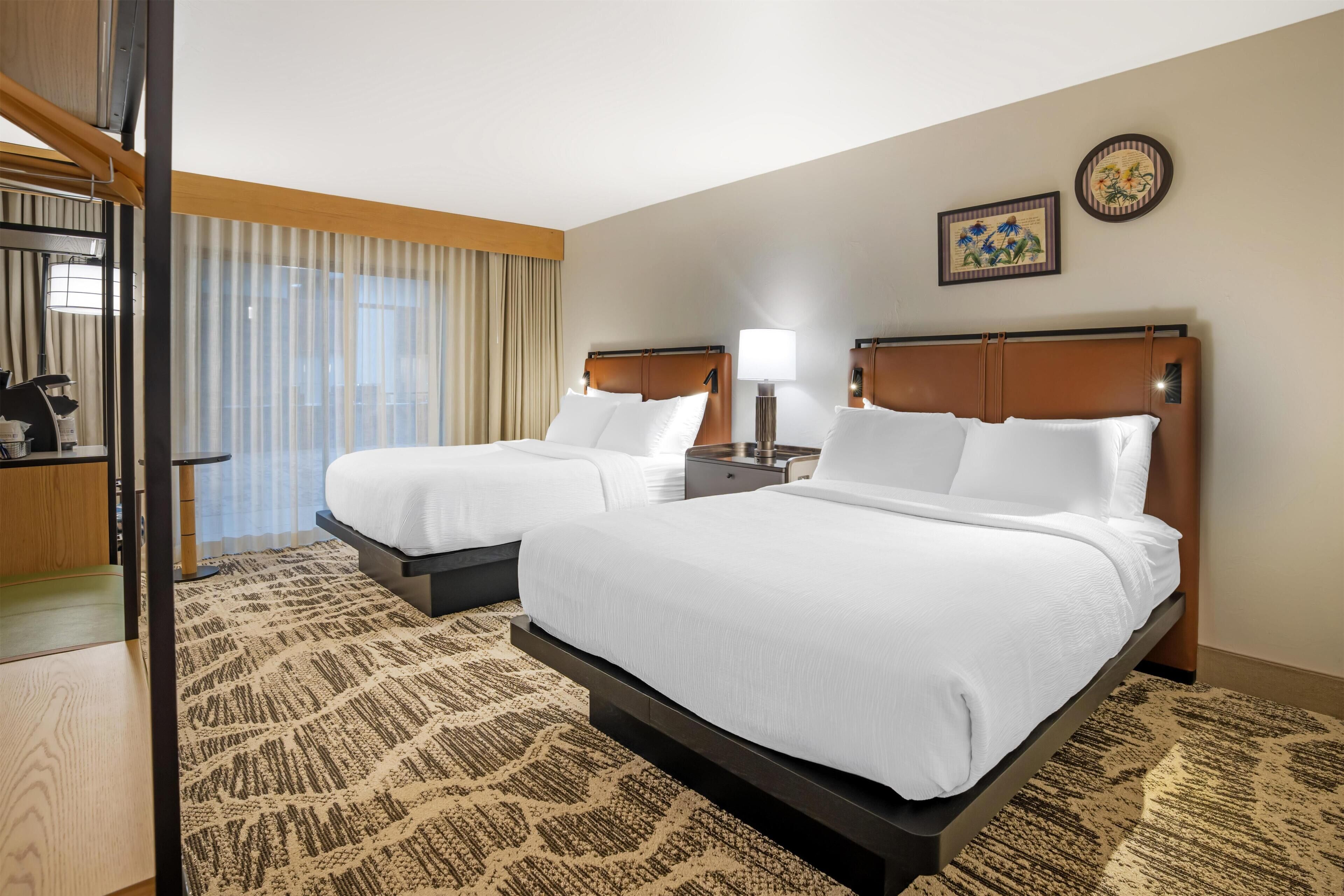 A hotel room with two beds, a desk, a chair, a lamp, and a window with curtains.