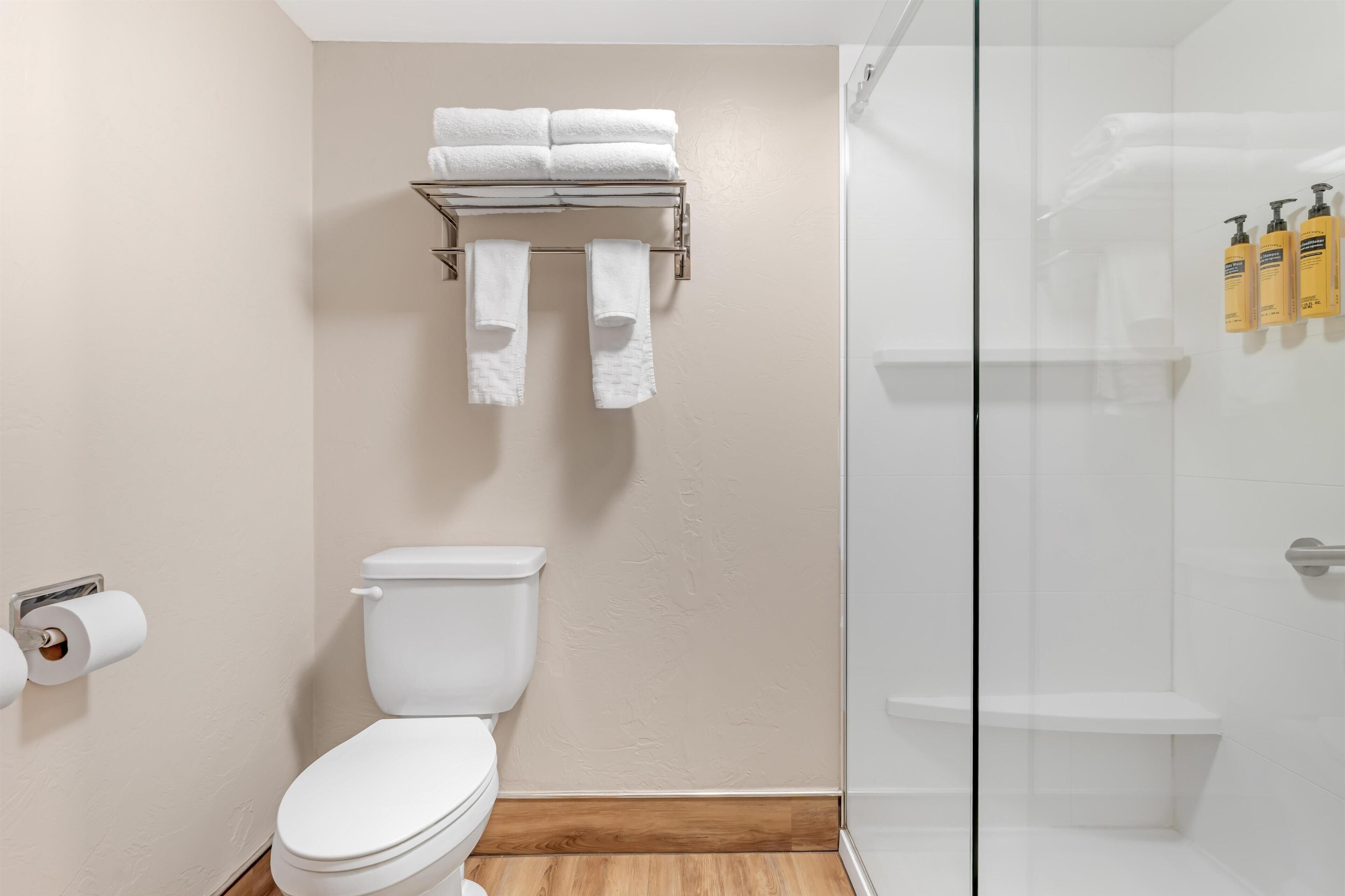 Et badeværelse med toilet, et håndklædestativ med håndklæder, en glas brusekabine og toiletartikler på væggen.