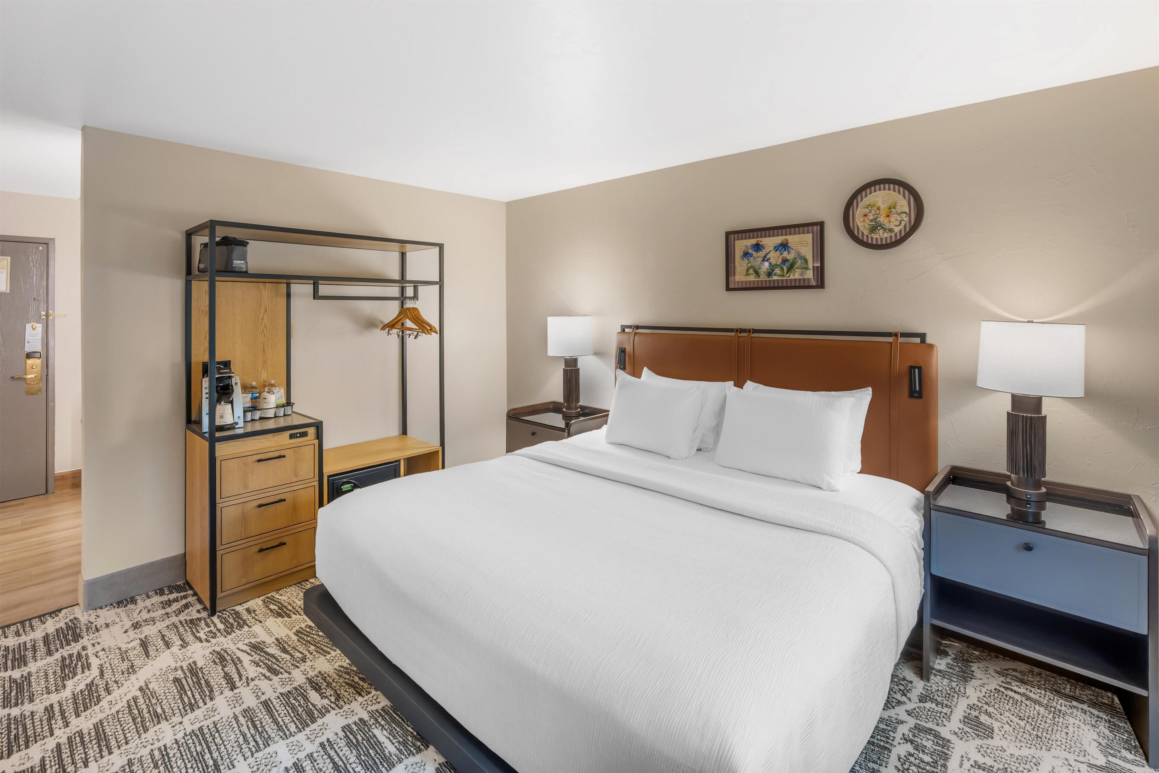 A hotel room with a large bed, a wooden dresser, two bedside tables with lamps, a mirror, and a wall-mounted picture.