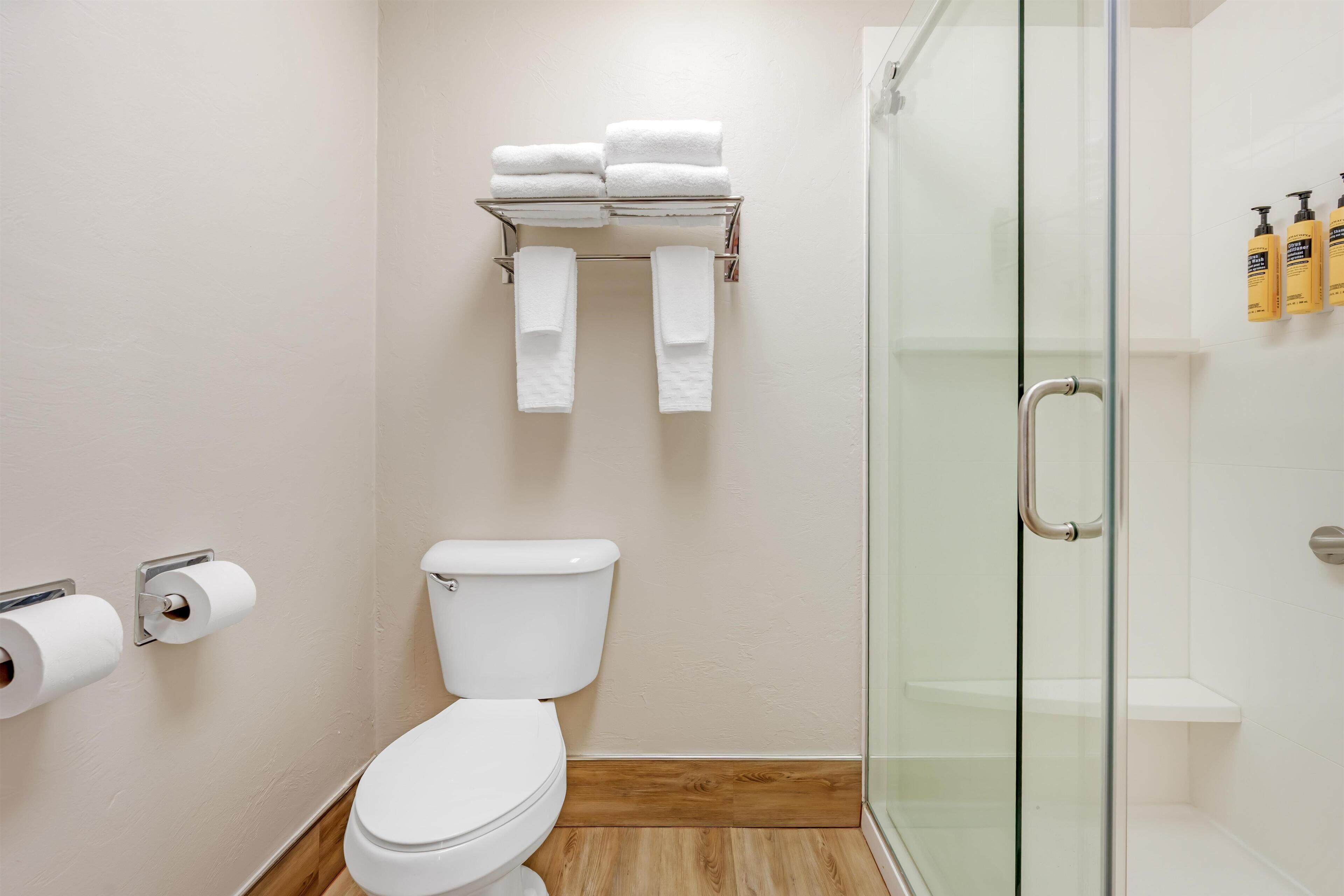 A bathroom with a toilet, a glass shower enclosure, a towel rack with folded towels, and toiletries on the wall.
