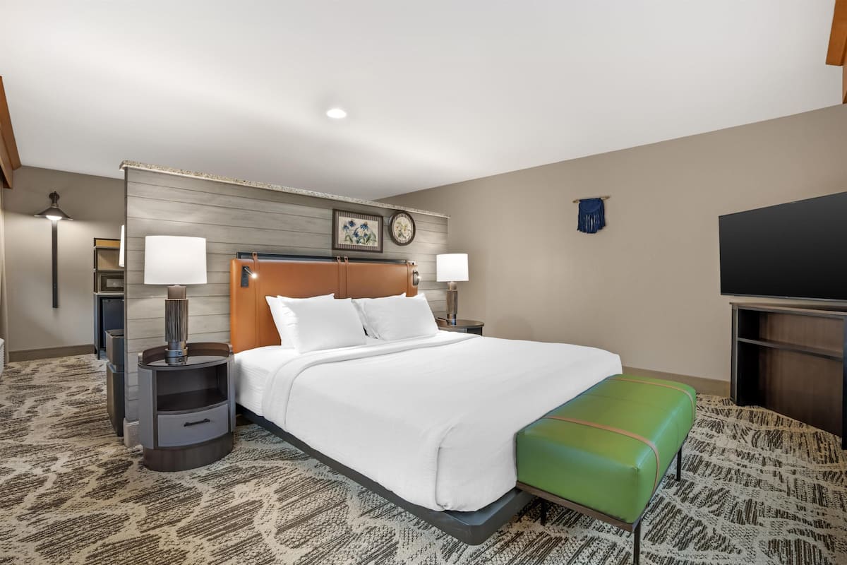 A hotel room with a bed, two bedside tables, a green bench, a TV, and a wall-mounted clock.