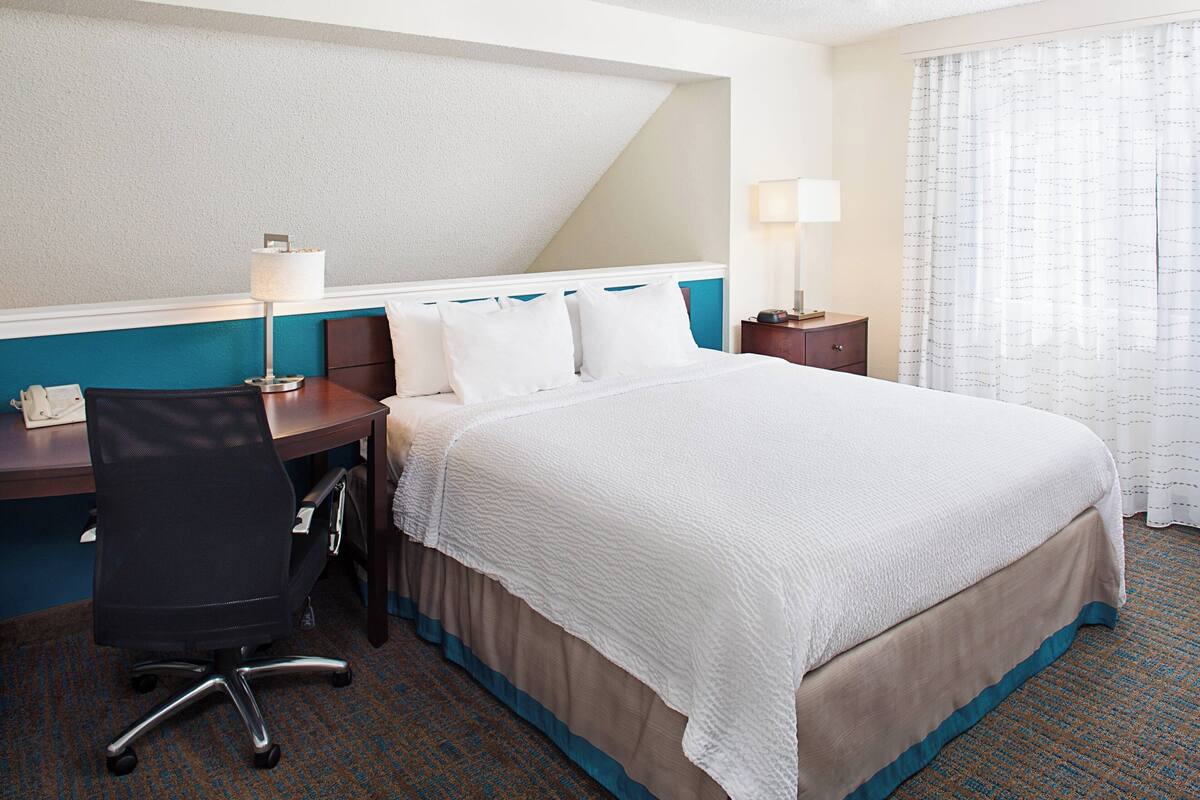 A hotel room with a bed, desk, chair, lamp, and curtains.