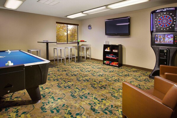 A room with a pool table, bar stools, a dartboard, and a television on the wall.