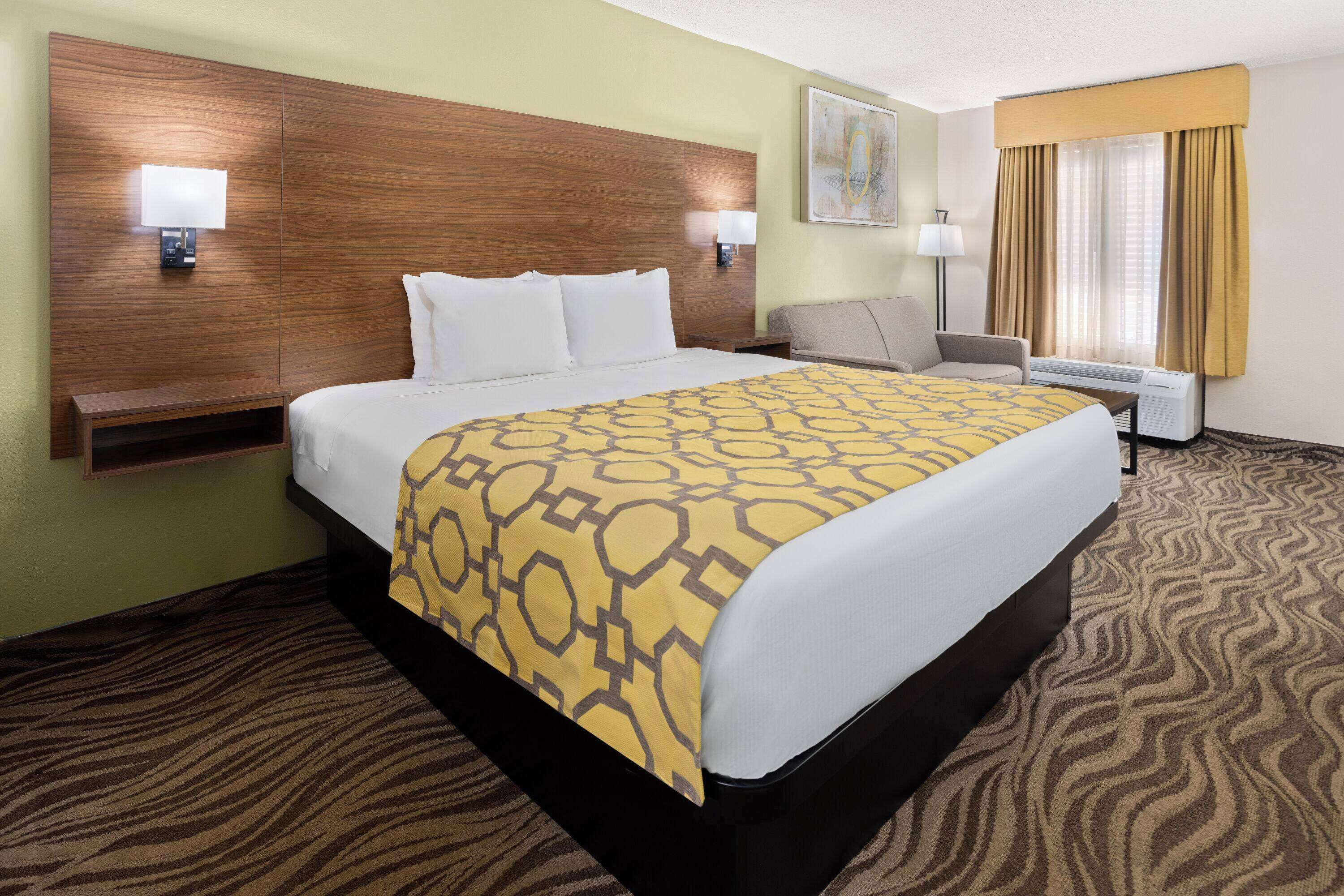 A hotel room with a large bed, a desk, a chair, a nightstand, and a window with curtains.