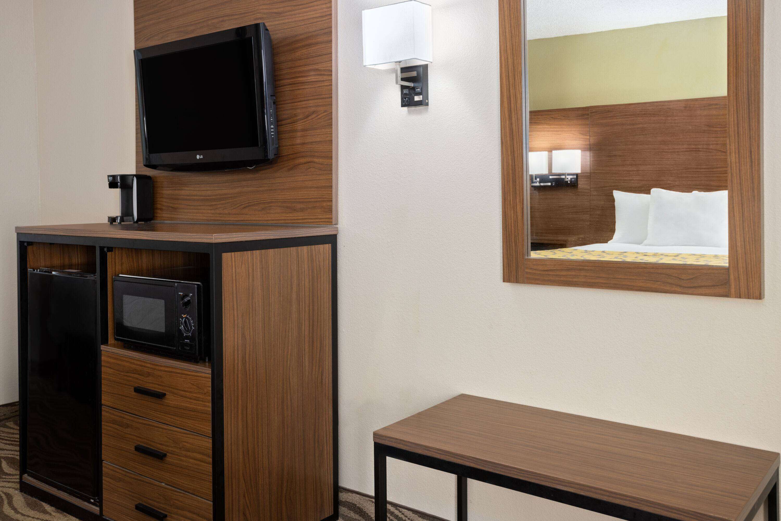 A hotel room with a wooden cabinet, a flat-screen TV, a microwave, a bedside table, and a large mirror reflecting the bed.