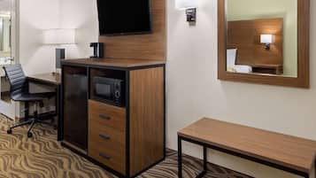 Habitación de hotel con un escritorio de madera, un televisor de pantalla plana montado encima, una mesita de noche pequeña, una silla y un espejo en la pared.