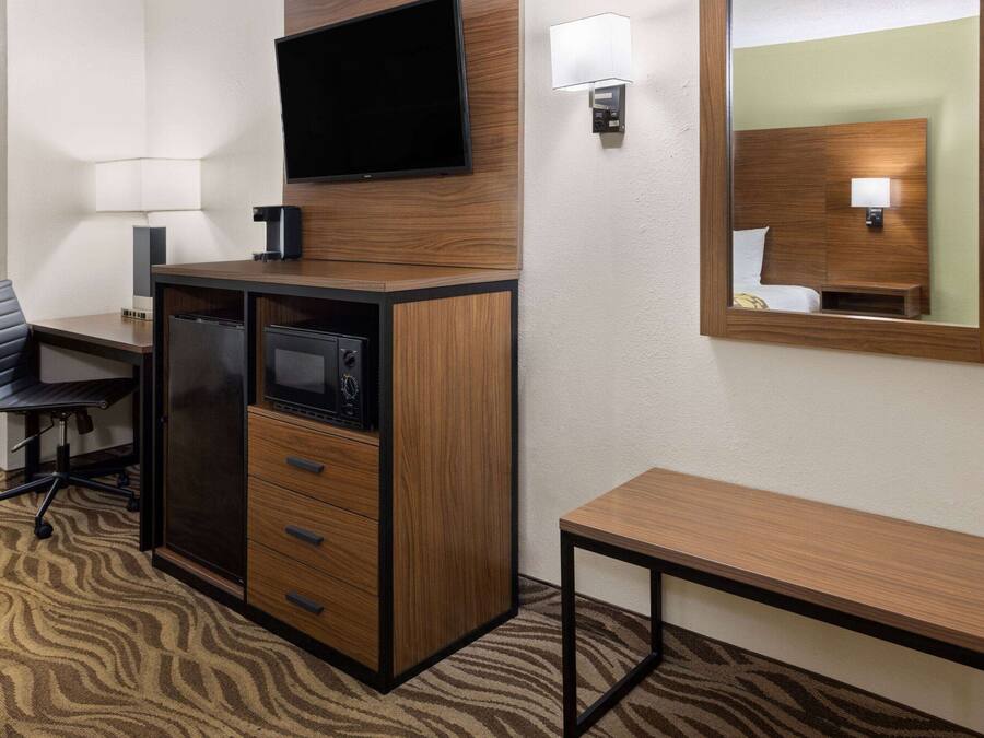 Habitación de hotel con un escritorio de madera, un televisor de pantalla plana montado encima, una mesita de noche pequeña, una silla y un espejo en la pared.