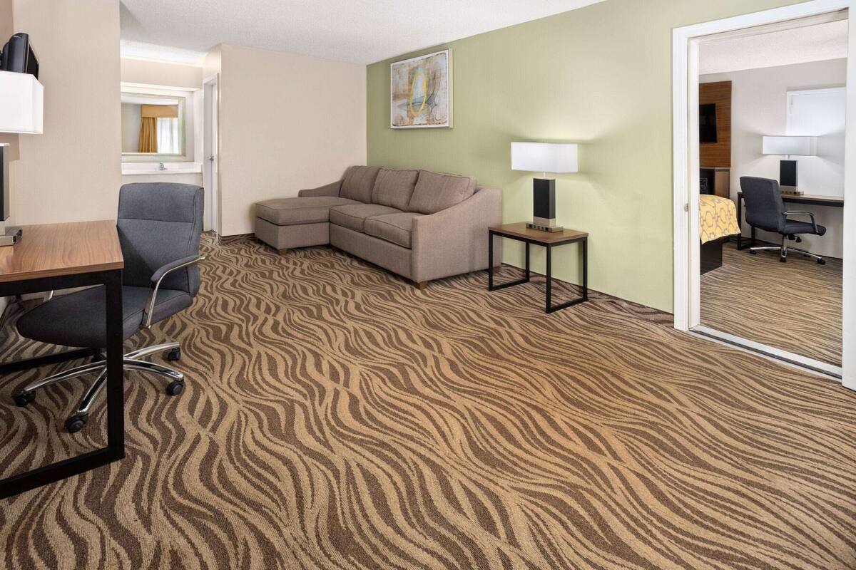 A hotel room with a sectional sofa, a desk with a chair, a small table with a lamp, and a bed in the adjacent room.