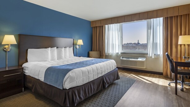 A hotel room with a large bed, two bedside lamps, a desk with a chair, a large window with curtains, and an air conditioning unit.