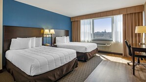 A hotel room with two beds, a desk, and a view of a cityscape.