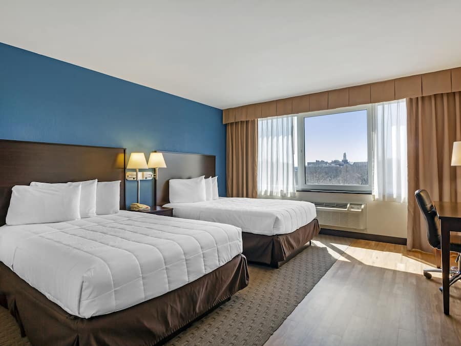 A hotel room with two beds, a desk, and a view of a cityscape.