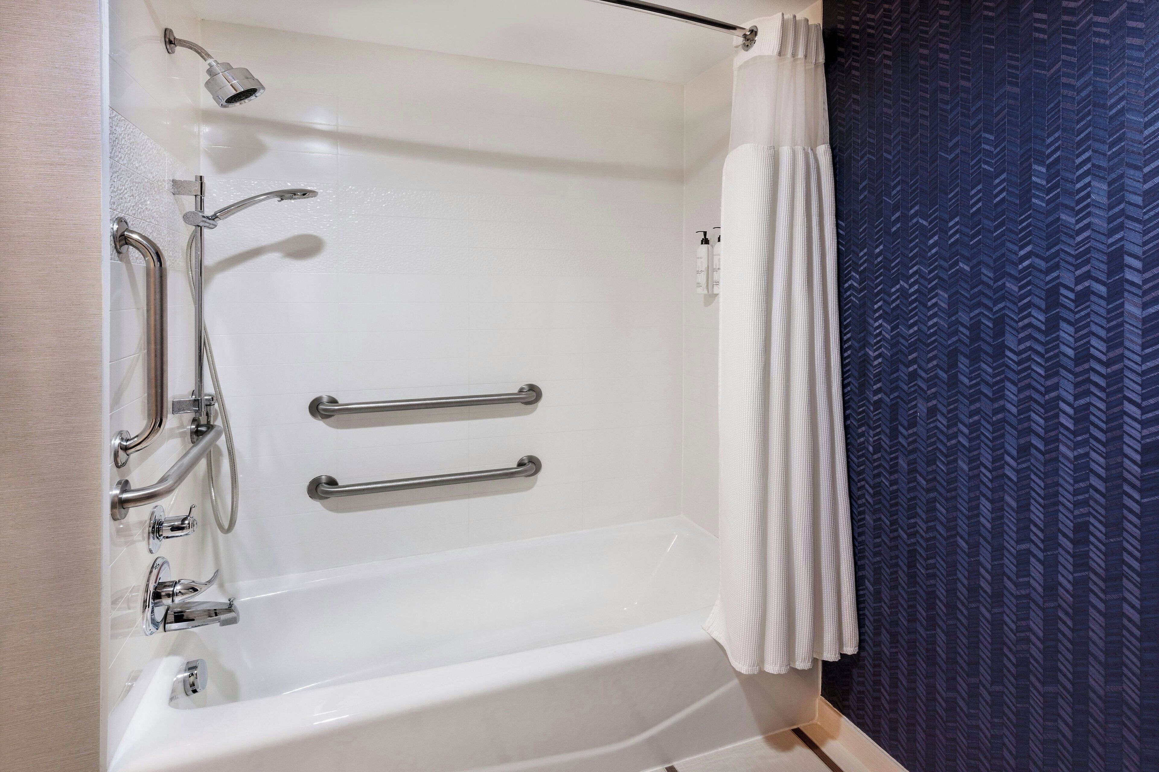 Un baño moderno con una bañera blanca, barras de apoyo, un cabezal de ducha manual y una cortina de ducha azul estampada.