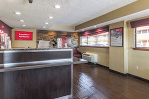 A hotel reception area with a counter, a sign indicating free Wi-Fi in all rooms, a Red Roof sign, and a window with a view of the outside. - Red Roof Inn Detroit - Troy (Troy)