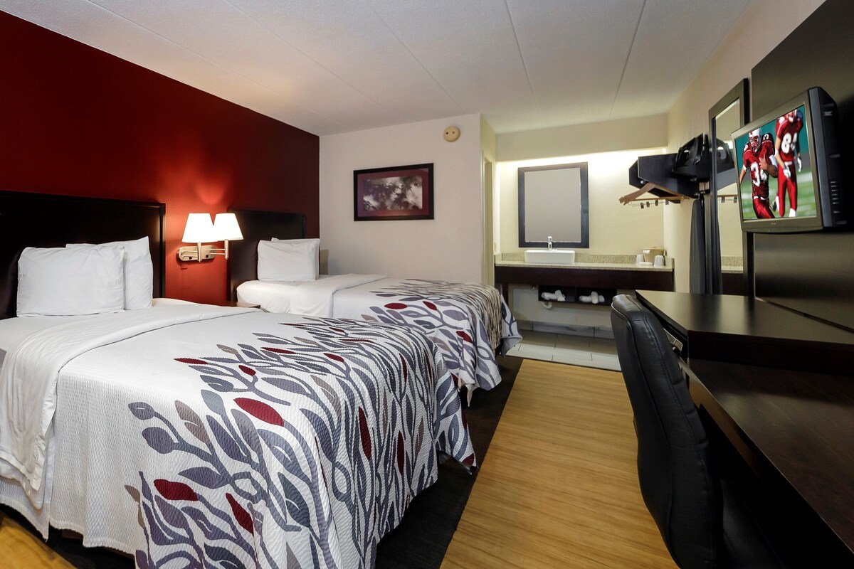 A hotel room with two beds, a desk, a TV, a mirror, and a bathroom.
