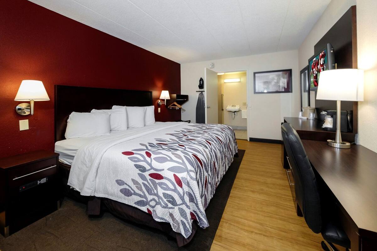 A hotel room with a bed, bedside lamps, a desk, a chair, a TV, and a bathroom visible in the background.