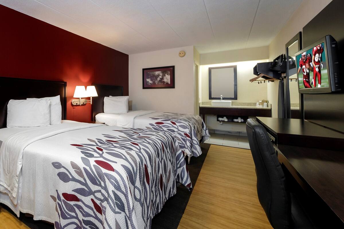 A hotel room with two beds, a desk, a TV, a mirror, and a bathroom.